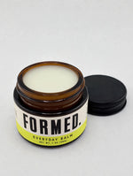 An open amber glass jar of Formed Everyday Balm with a black screw cap resting beside it on a light gray background.
