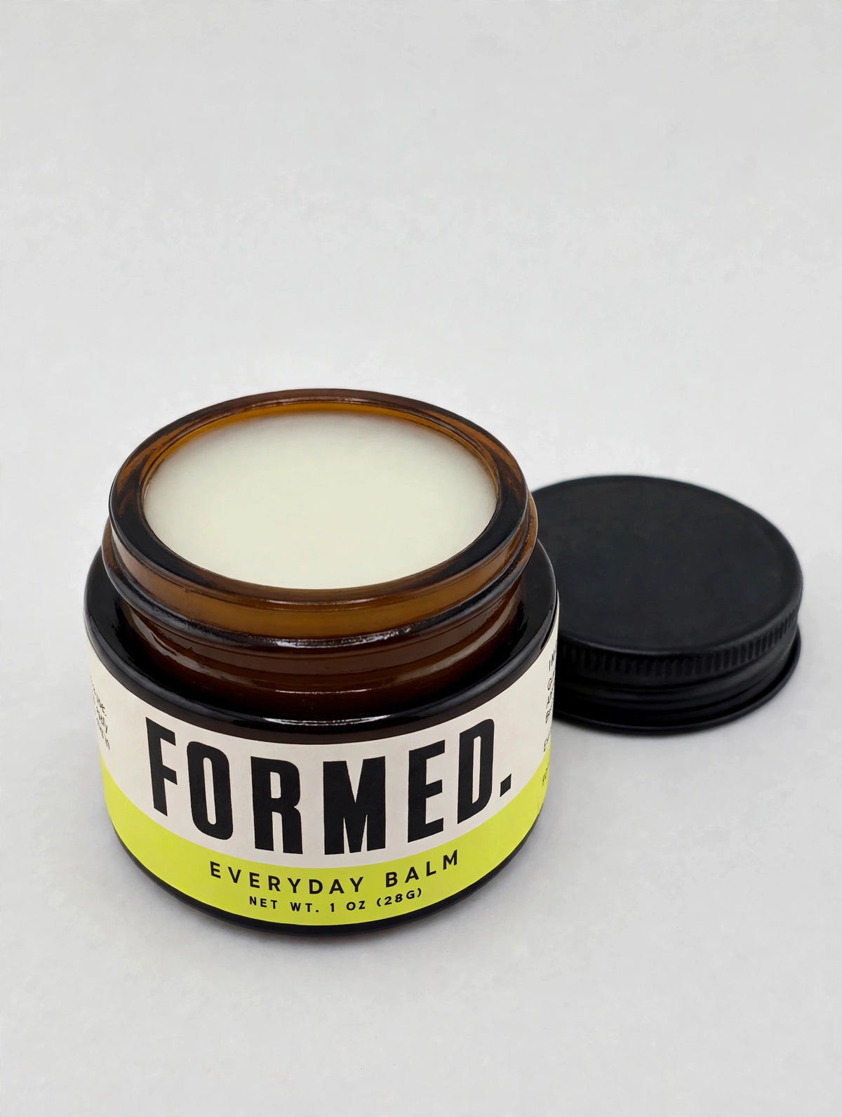 An open amber glass jar of Formed Everyday Balm with a black screw cap resting beside it on a light gray background.